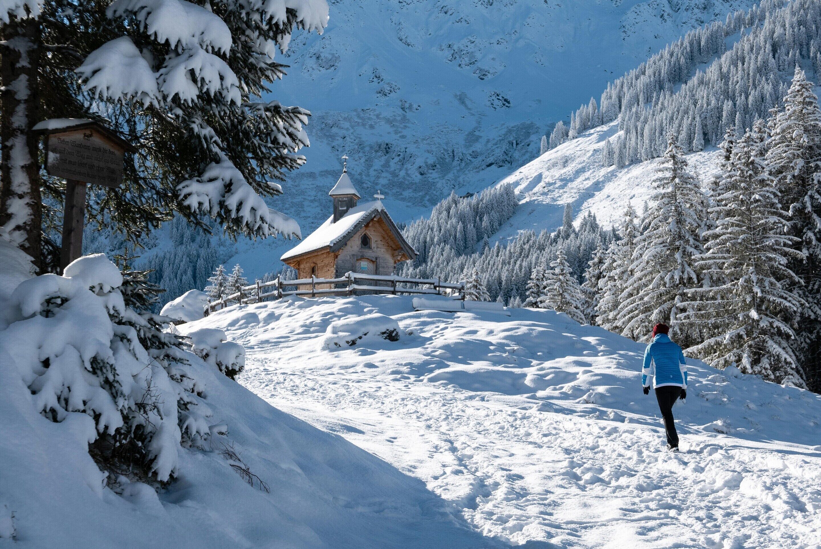 Un sentier de montagne enneigé menant à un chalet en bois doté d’un clocher, entouré d’arbres chargés de neige.