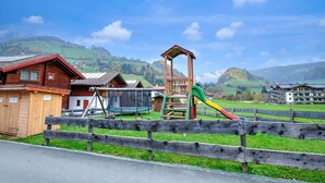 A playground with a slide and climbing structure, surrounded by wooden buildings and a fenced area. - Feriendorf Wildschonau-near ski Area (Wildschönau)