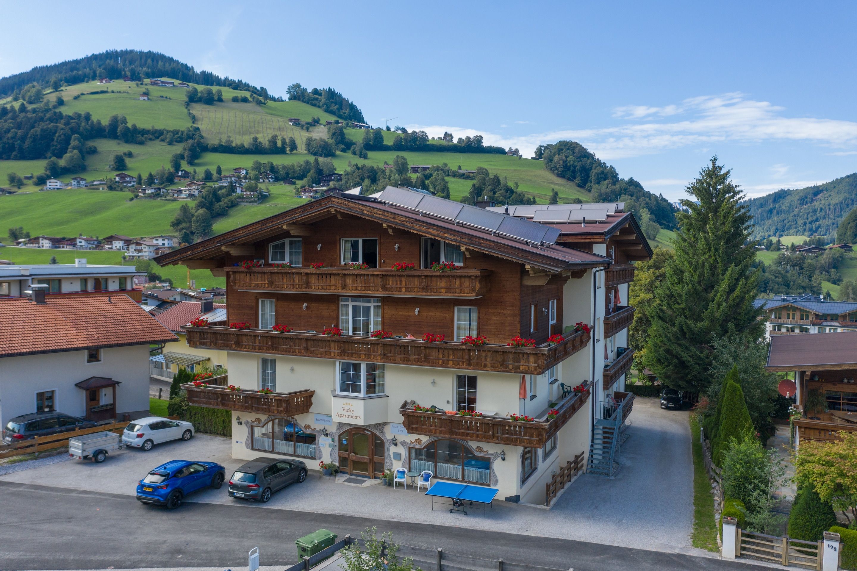 Un edificio de varios pisos con estilo arquitectónico tradicional, que cuenta con balcones con macetas de flores, una zona de recepción y un estacionamiento con coches.