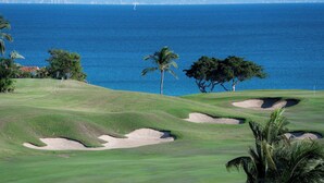 Campo de golf con búnkeres de arena y palmeras, con vistas a una ciudad costera.