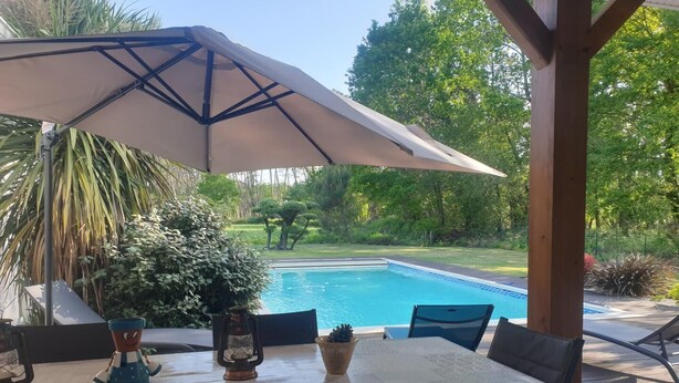 En terrasse med et bord og stole, en stor parasol og udsigt til en swimmingpool.