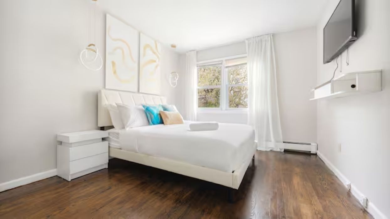A modern bedroom with a large bed, white walls, and a window with curtains.