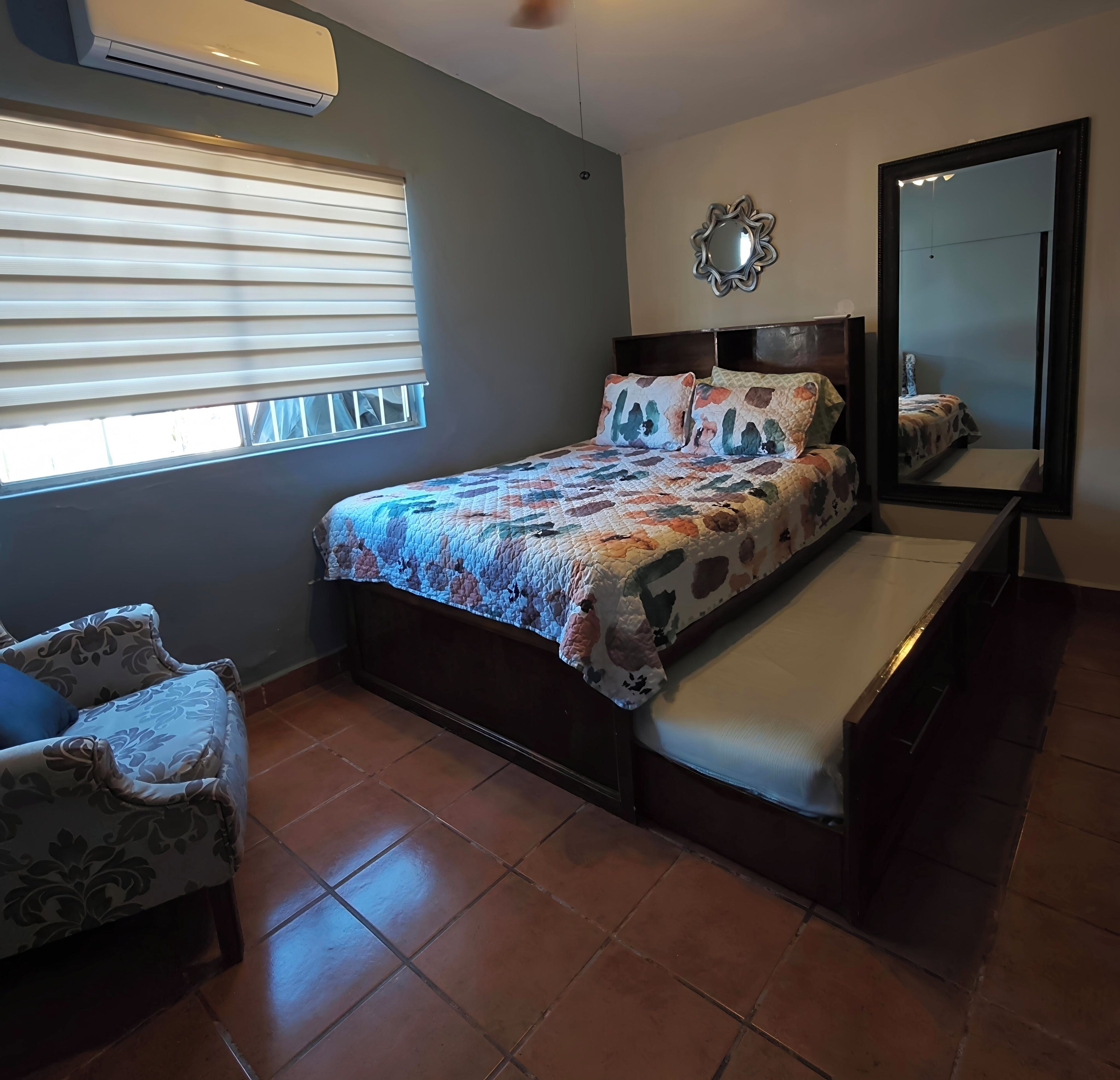 A bedroom with a bed, a chair, a window with blinds, a wall-mounted mirror, and a ceiling fan.
