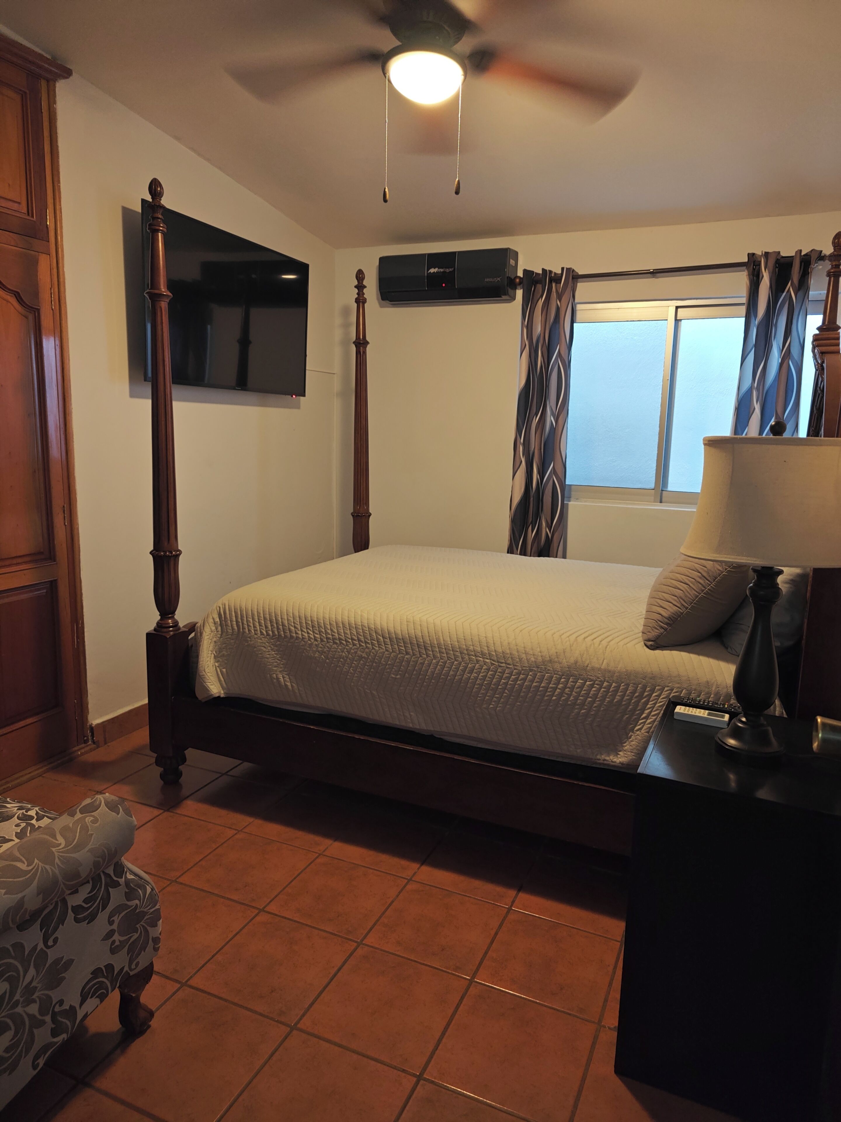 A bedroom with a four-poster bed, a ceiling fan, a TV mounted on the wall, and a window with curtains.