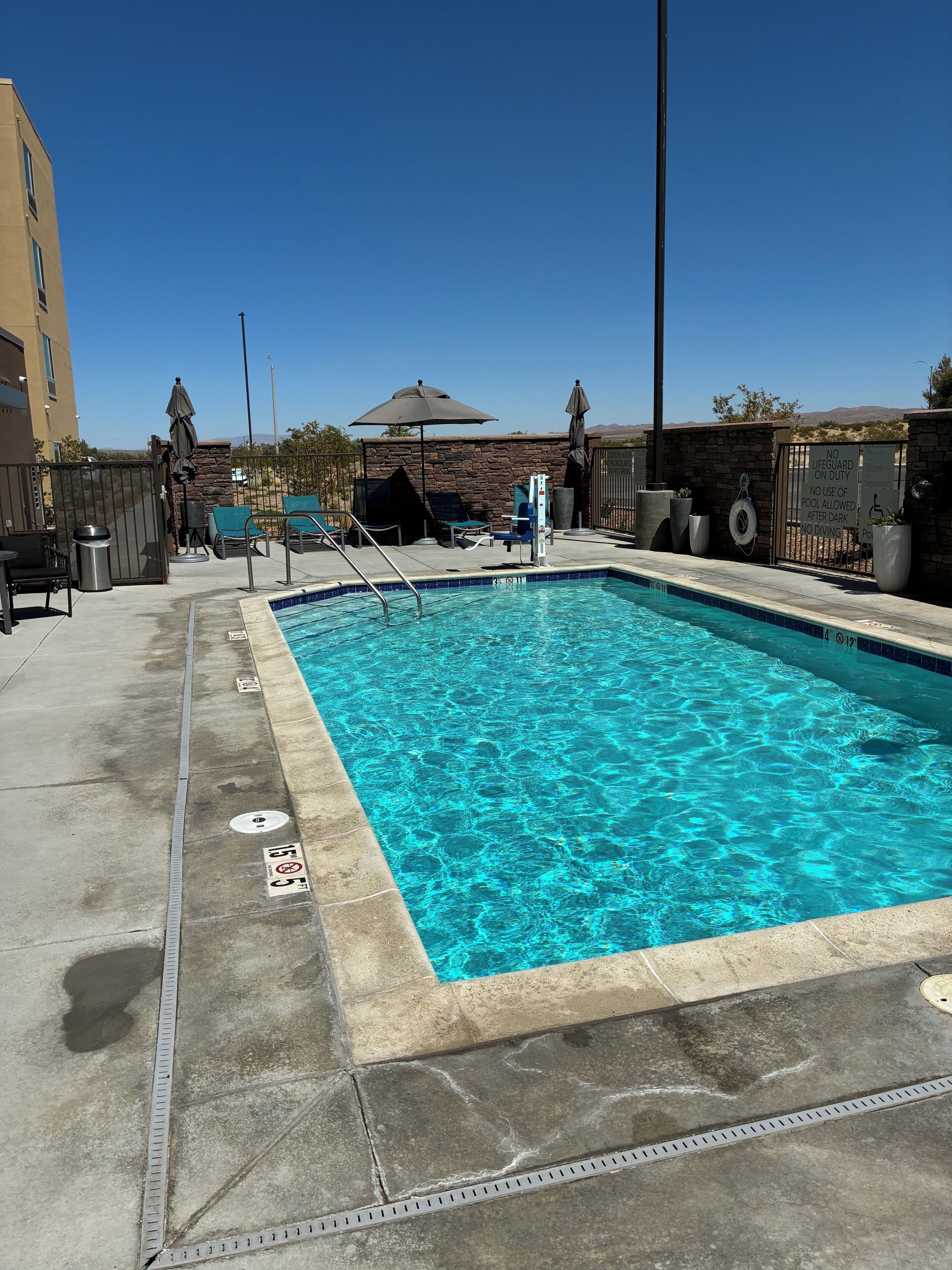 A rectangular swimming pool with clear blue water, surrounded by a concrete deck with lounge chairs and umbrellas.