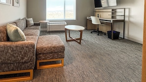 A furnished room with a sofa, a small round table, a desk with a chair, a TV, and a large window. - Towneplace Suites By Marriott Ridgecrest (Ridgecrest)