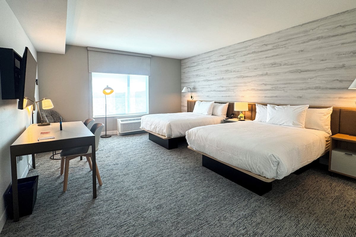 A hotel room with two beds, a desk, a chair, a TV, and a large window.