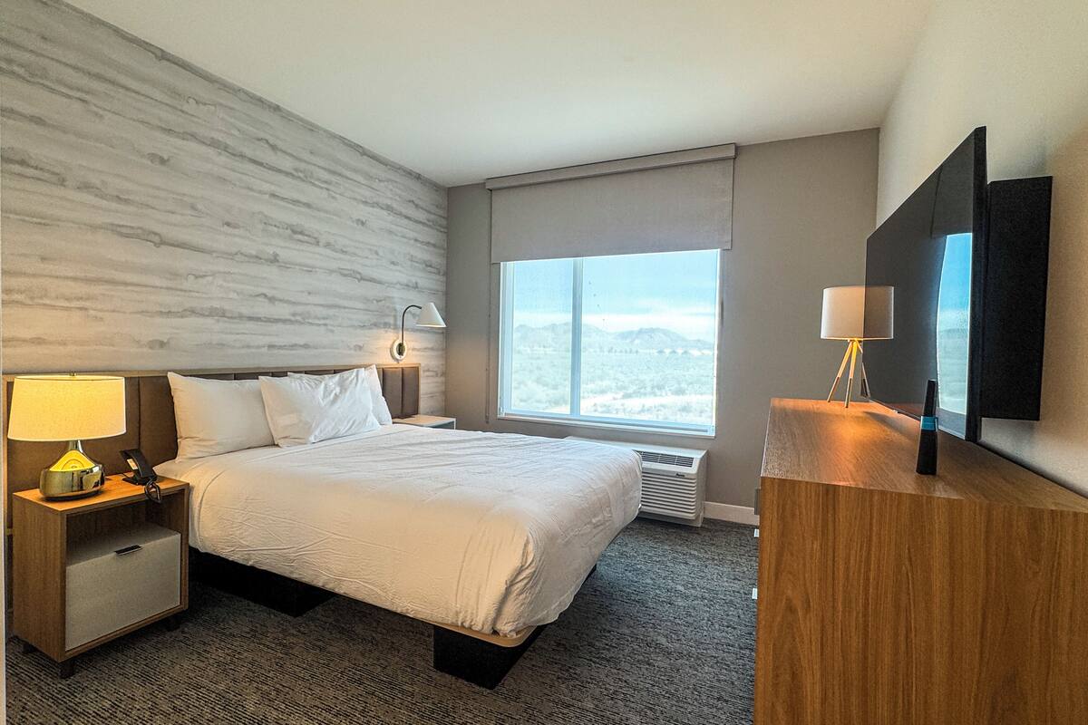 A hotel room with a large bed, a nightstand with a lamp, a desk with a lamp, a large window with a view, and a flat-screen TV mounted on the wall.