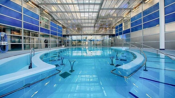 A large indoor swimming pool with clear blue water, surrounded by glass walls and a metal ceiling.