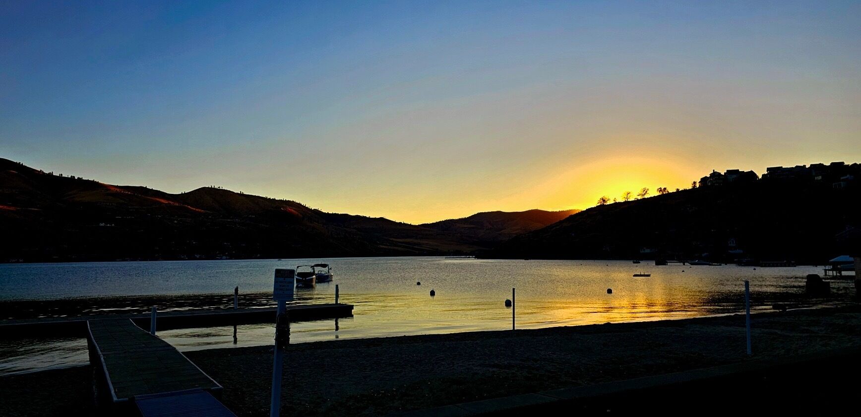 Atardecer sobre un lago tranquilo con un muelle y barcos, rodeado de colinas.