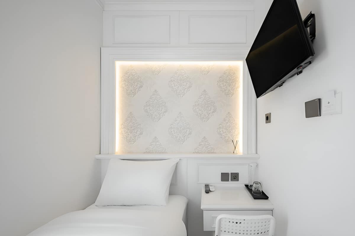 A modern hotel room with a neatly made bed, a bedside table with a lamp, a flat-screen TV mounted on the wall, and a decorative light panel above the bed.