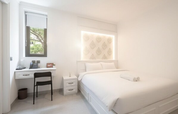 A bedroom with a bed, a desk with a chair, a nightstand, and a window with a view of trees. - Wyneth Hotel Airport (Tangerang)