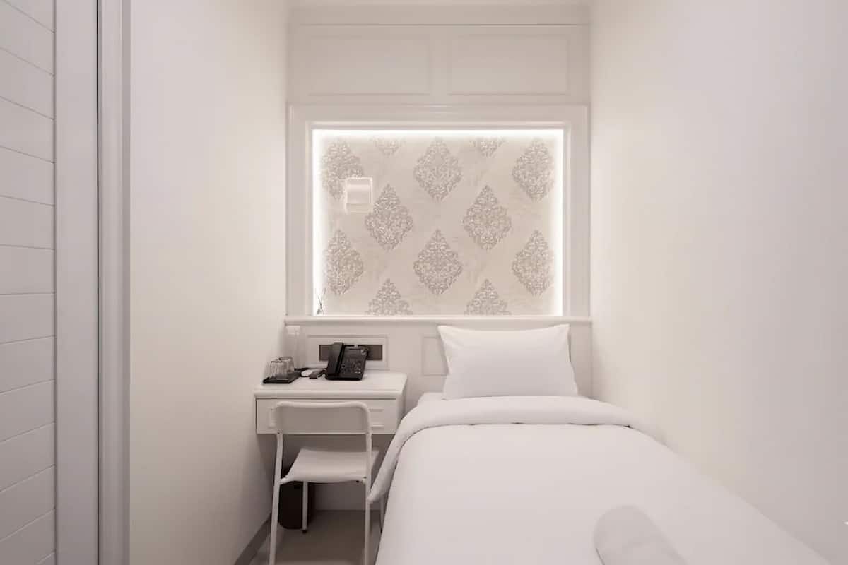 A small, white, single-bed room with a desk and chair, a window with patterned curtains, and a white wall.