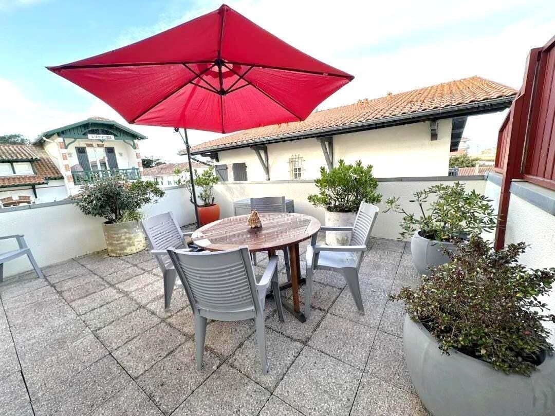A patio with a round table, four chairs, a red umbrella, and potted plants.