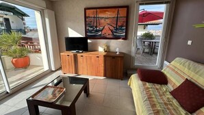Una sala de estar con sofá, mesa de centro, televisor y aparador de madera. Hay un cuadro en la pared y vistas al exterior a través de un ventanal.