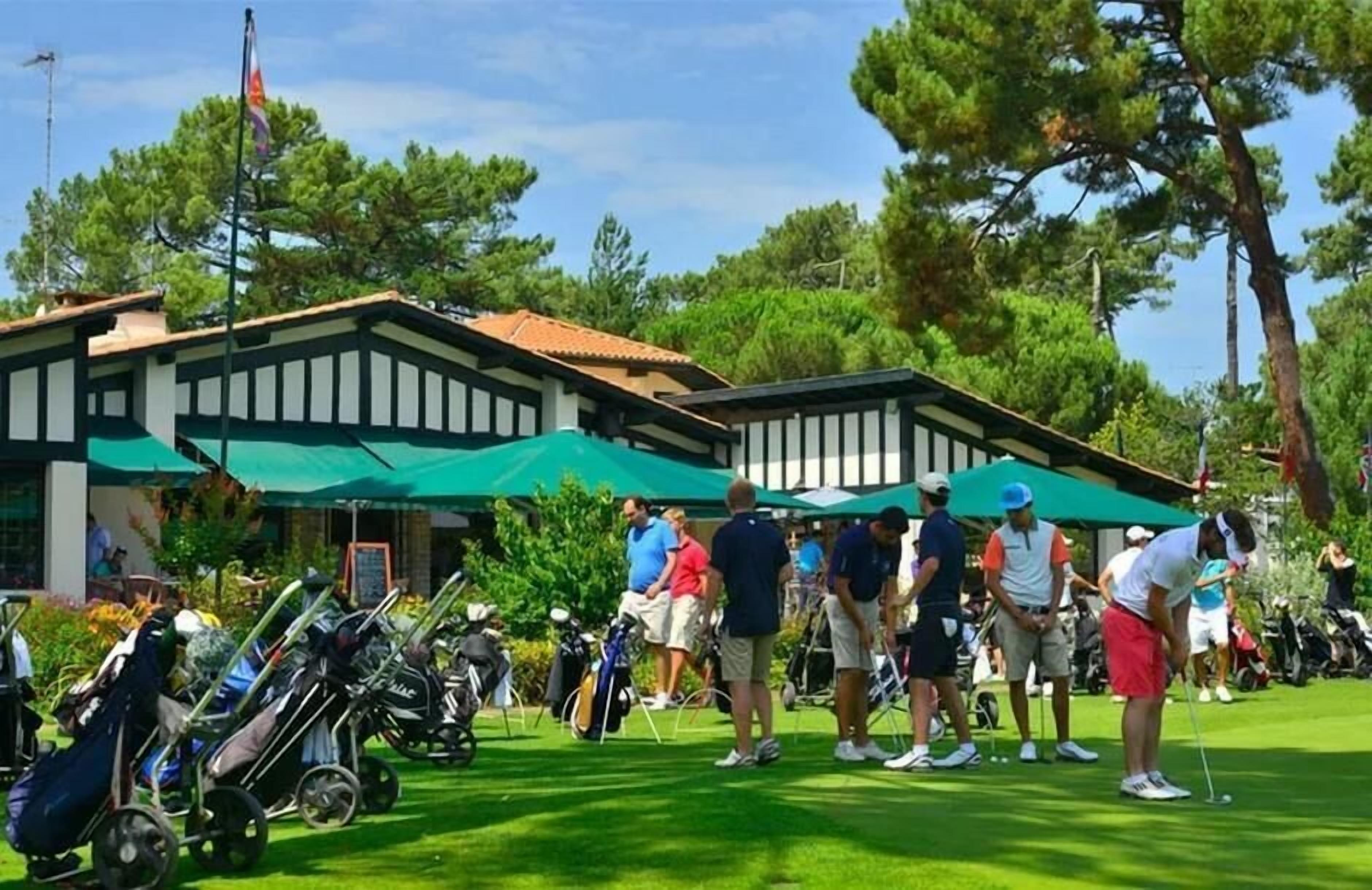 En golfbane med spillere og tilskuere, en bygning med grønt tag og en flagstang.