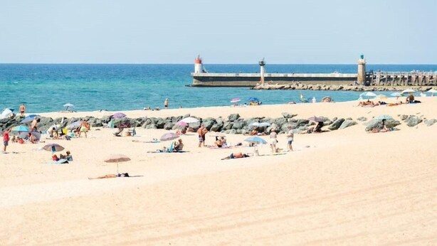 Una playa con gente tomando el sol y caminando, un muelle con faros y agua azul tranquila.