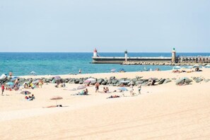 Uma praia com pessoas tomando sol e caminhando, um píer com faróis e água azul calma.