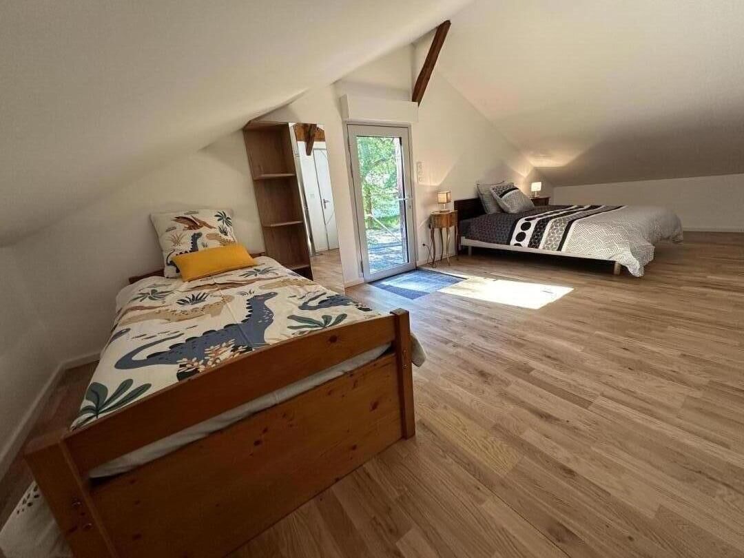 Une chambre avec un plafond en pente, un plancher en bois, un lit recouvert d’une couvre-lit à motifs floraux, une table de chevet avec une lampe, et une vue à travers une porte vitrée.