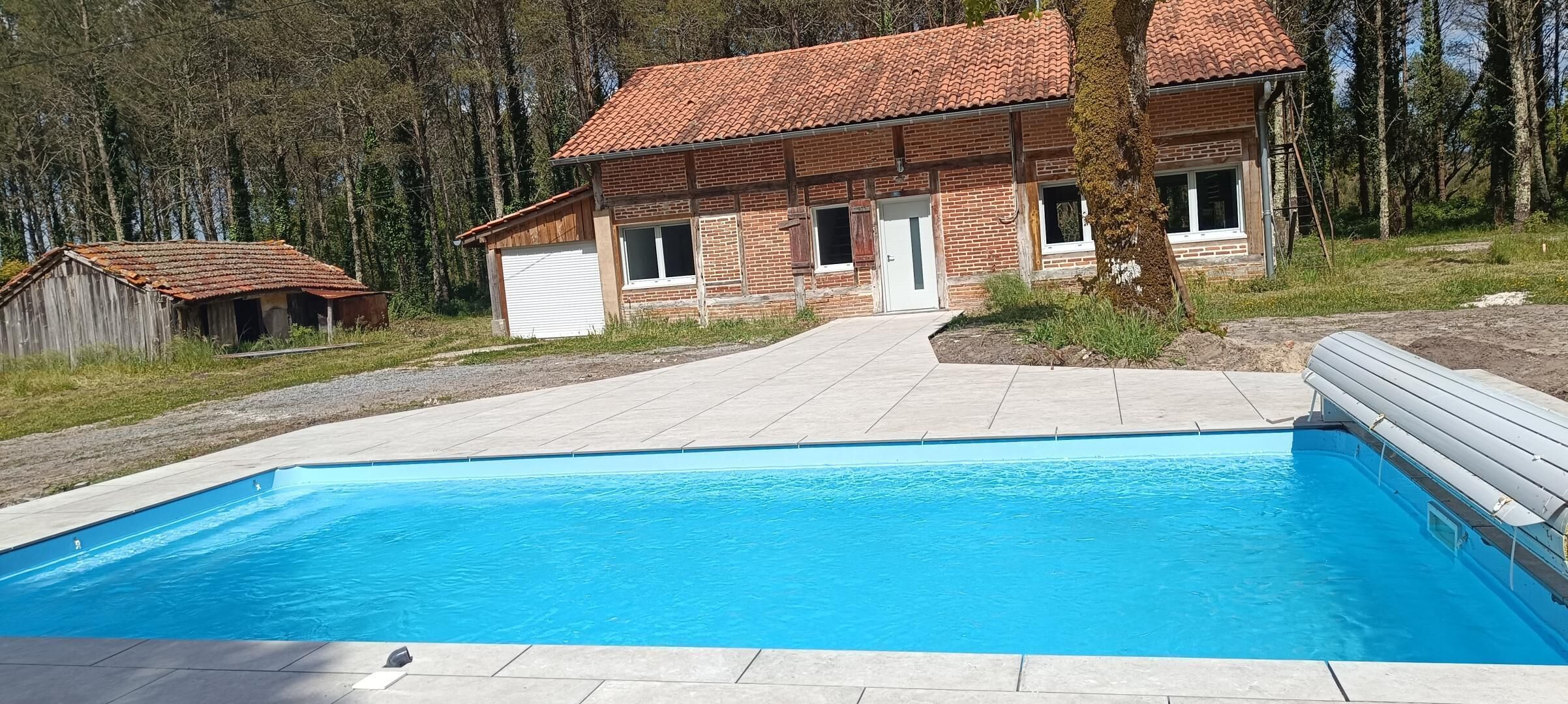 A swimming pool with a tiled deck, a brick house with a red-tiled roof, and a wooden shed in a wooded area.