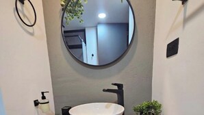 A bathroom with a round mirror, a white vessel sink, a black faucet, a soap dispenser, a small potted plant, and a towel rack. - Epic Suite 209 Deluxe Aeropuerto (Pereira)