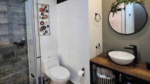 A modern bathroom with a glass shower enclosure, a white toilet, a wooden vanity with a round mirror, and a stone wall. - Epic Suite 209 Deluxe Aeropuerto (Pereira)