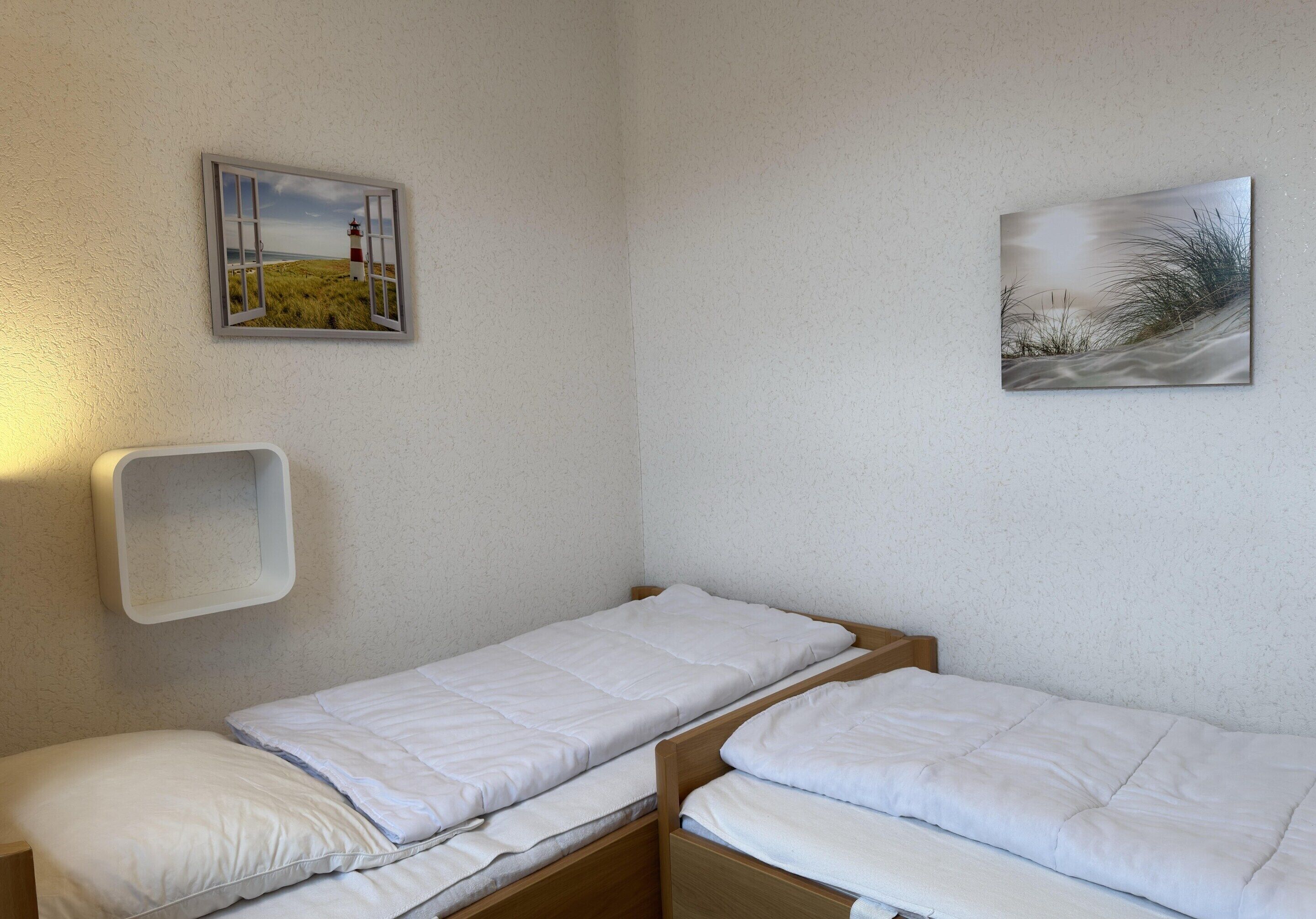A small, single-room accommodation with two wooden beds, a white wall, and two framed pictures.