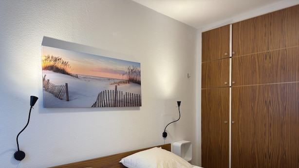 A hotel room with a bed, a wooden wardrobe, a wall-mounted picture, and two wall-mounted lamps. - Bungalow mit Garten in Dornumersiel (Dornum)