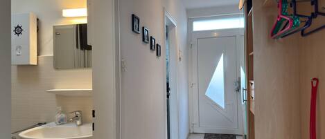 Corredor de hotel com um banheiro à esquerda, uma porta com janela triangular e um porta-roupas com objetos coloridos.