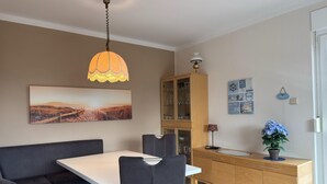 Ein Essbereich mit einem weißen Tisch, schwarzen Stühlen, einem Holzschrank, einer Hängelampe und einem Gemälde einer Strandlandschaft.