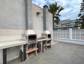Eine Dachterrasse mit eingebautem Grill und Schornstein, einem weißen Zaun und einem Gebäude im Hintergrund.