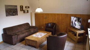 Ein Wohnzimmer mit braunem Sofa, einem Couchtisch aus Holz, einem Ledersessel, einem Fernseher auf einem Holzboden und einer Pendelleuchte.