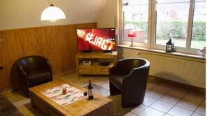 Ein Wohnzimmer mit einem Holzboden-TV-Schrank, einem Flachbildfernseher, der 'SEJAGT' zeigt, zwei Ledersesseln, einem Couchtisch mit Untersetzer, einer Weinflasche und einem Glas.