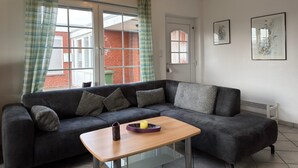 A modern living room with a dark grey sofa, a small wooden table, and a view of a brick building through large windows. - Bungalow in Strandnaehe, Dornumersiel (Dornum)