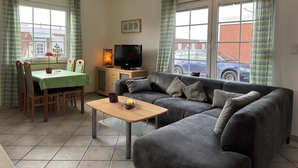 A living room with a sofa, a table, a TV, and a window with curtains. - Bungalow in Strandnaehe, Dornumersiel (Dornum)