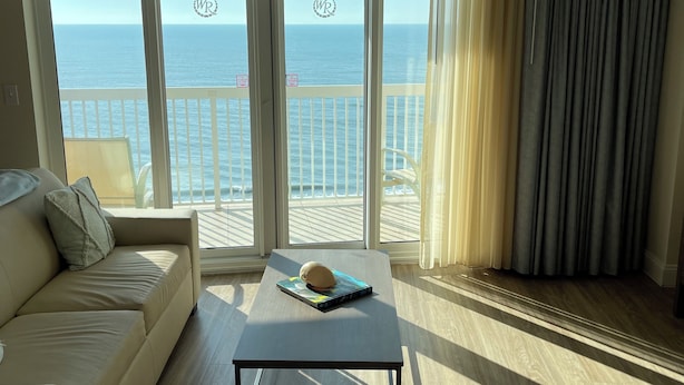 Un salon comprenant un canapé, une table basse et un ventilateur de plafond. De grandes fenêtres offrent une vue sur l’océan.