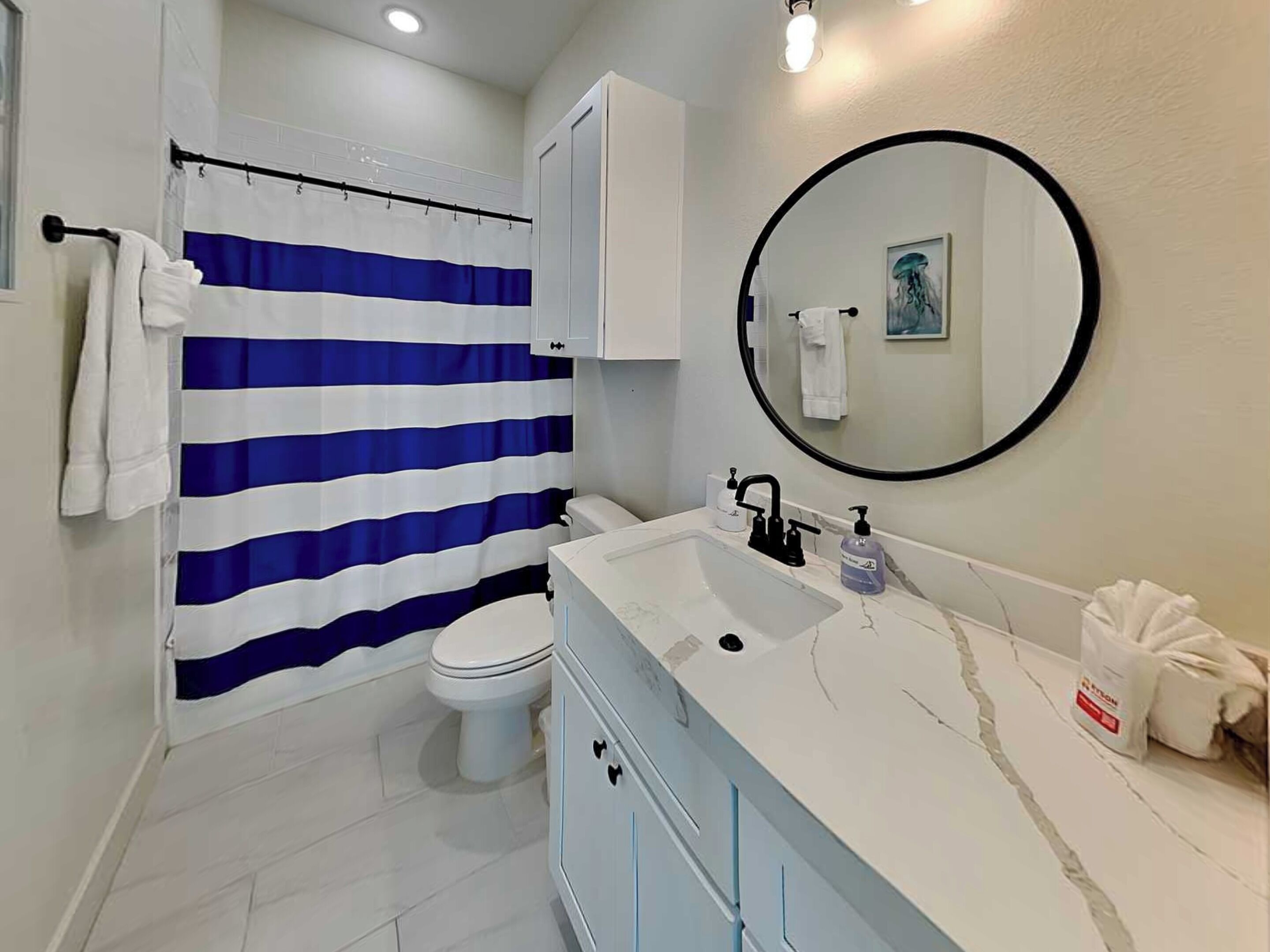 Un baño con un lavamanos blanco, un inodoro, una cortina de ducha azul y blanca a rayas, y un espejo redondo en la pared.