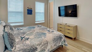 Un dormitorio con una cama, dos ventanas con persianas, un aparador, un televisor y un ventilador de techo.