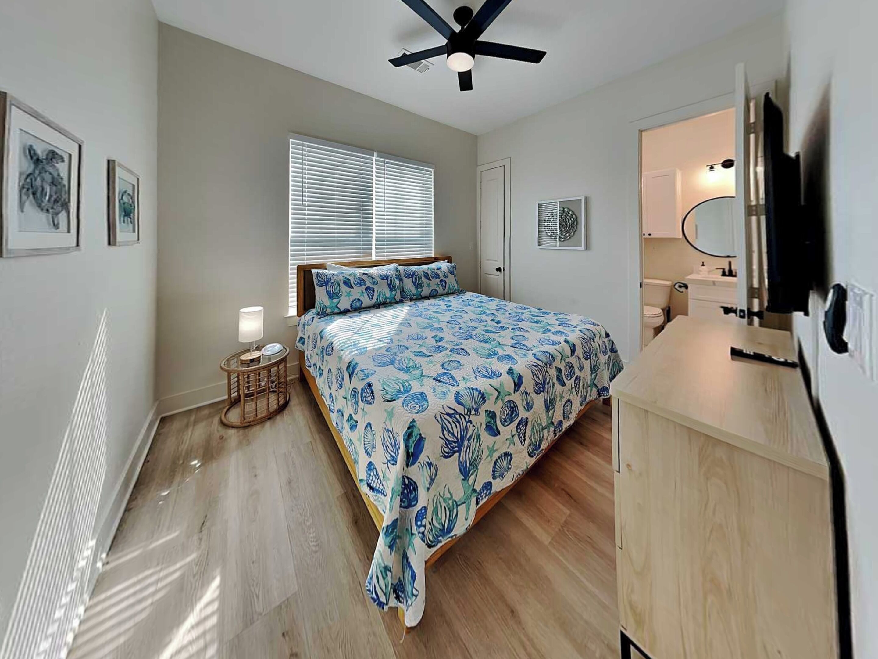 A bedroom with a bed, a ceiling fan, a window with blinds, a bedside table with a lamp, and a bathroom with a mirror and sink.