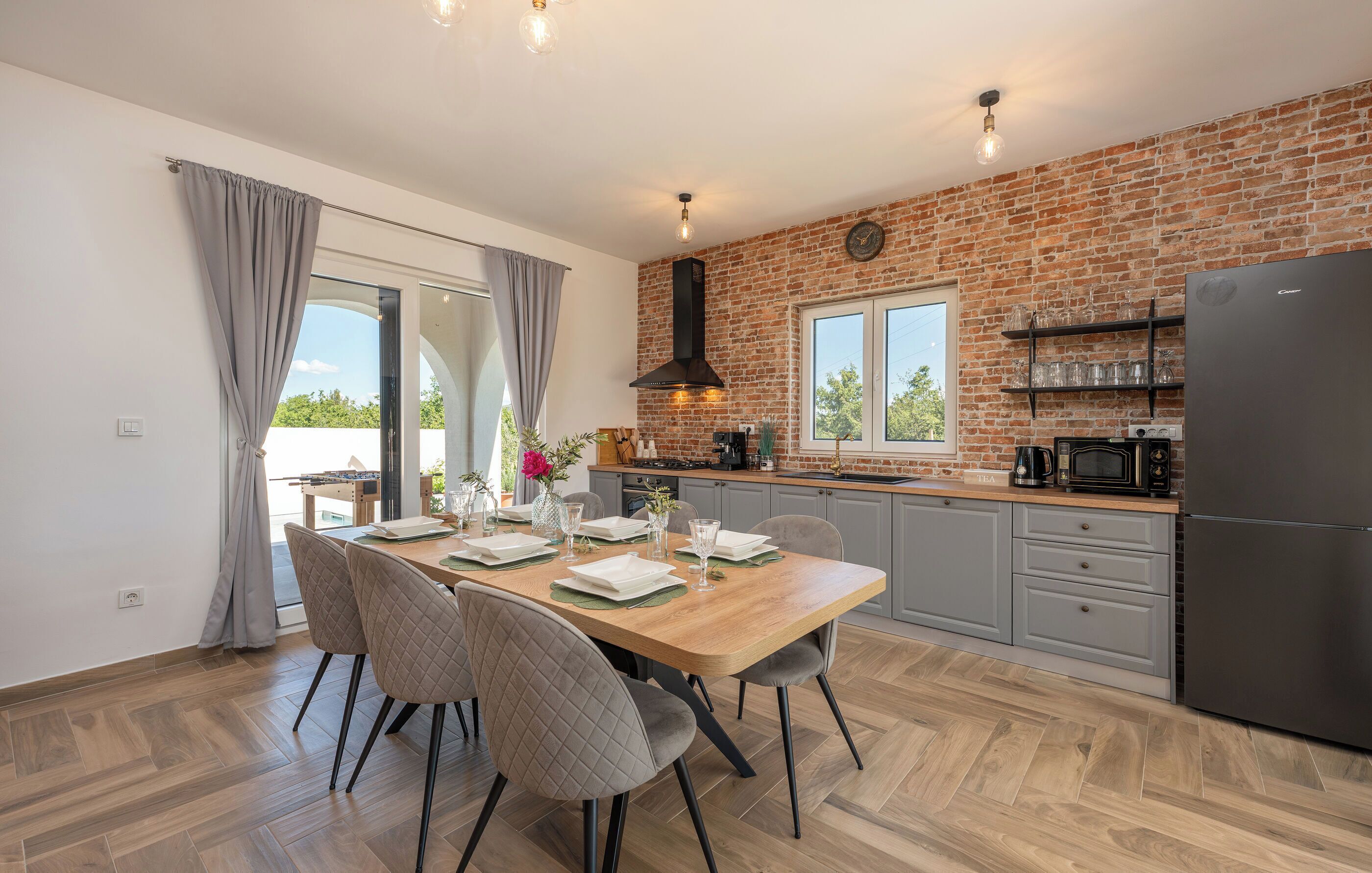 A modern kitchen with a brick wall, a dining table set for four, and a large refrigerator.