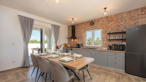 A modern kitchen with a brick wall, a dining table set for four, and a large refrigerator. - Villa Olivarum (Stankovci)