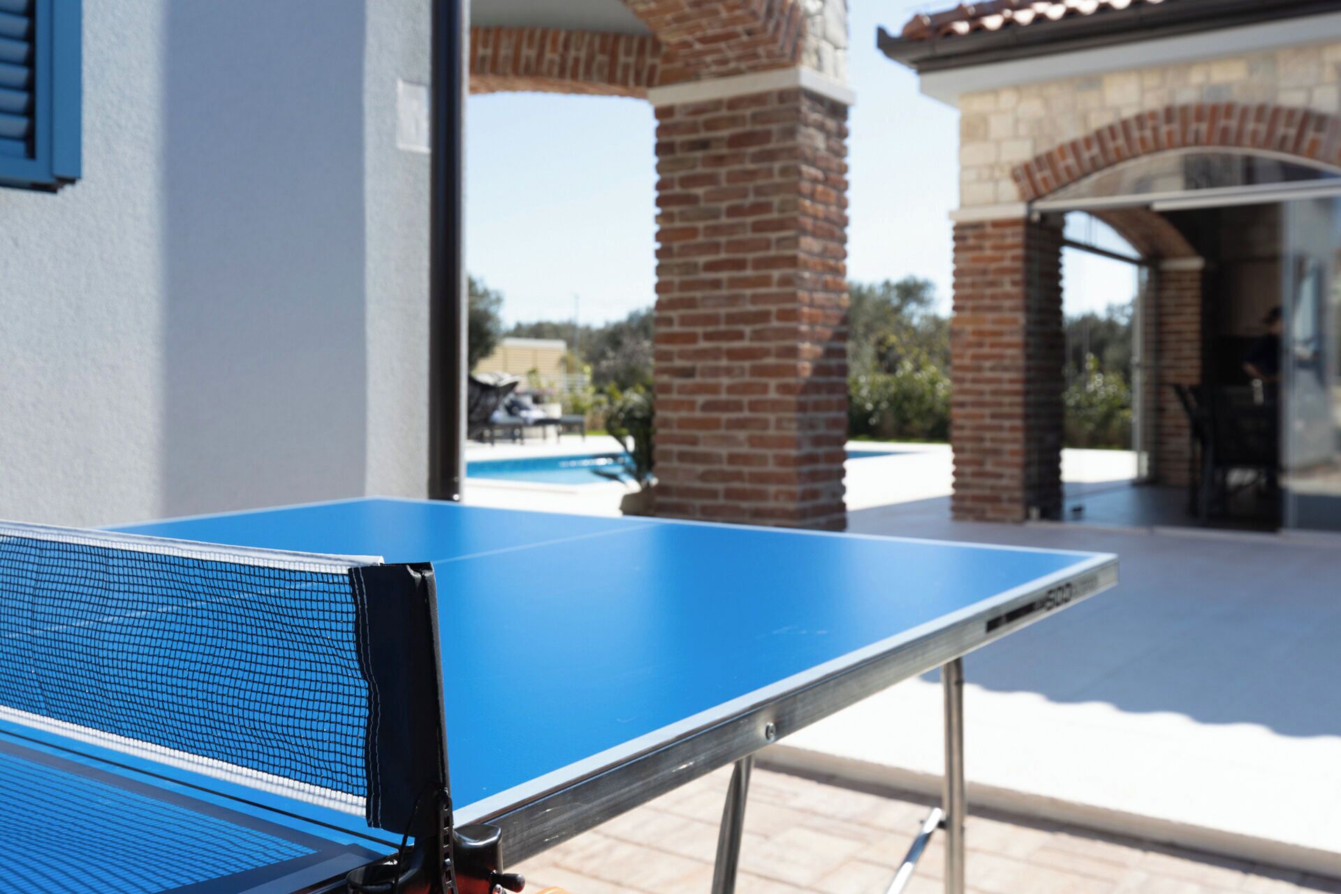 Une table de ping-pong bleue avec un filet, installée à l’extérieur, près d’un pilier en briques et d’une zone dédiée à la piscine.