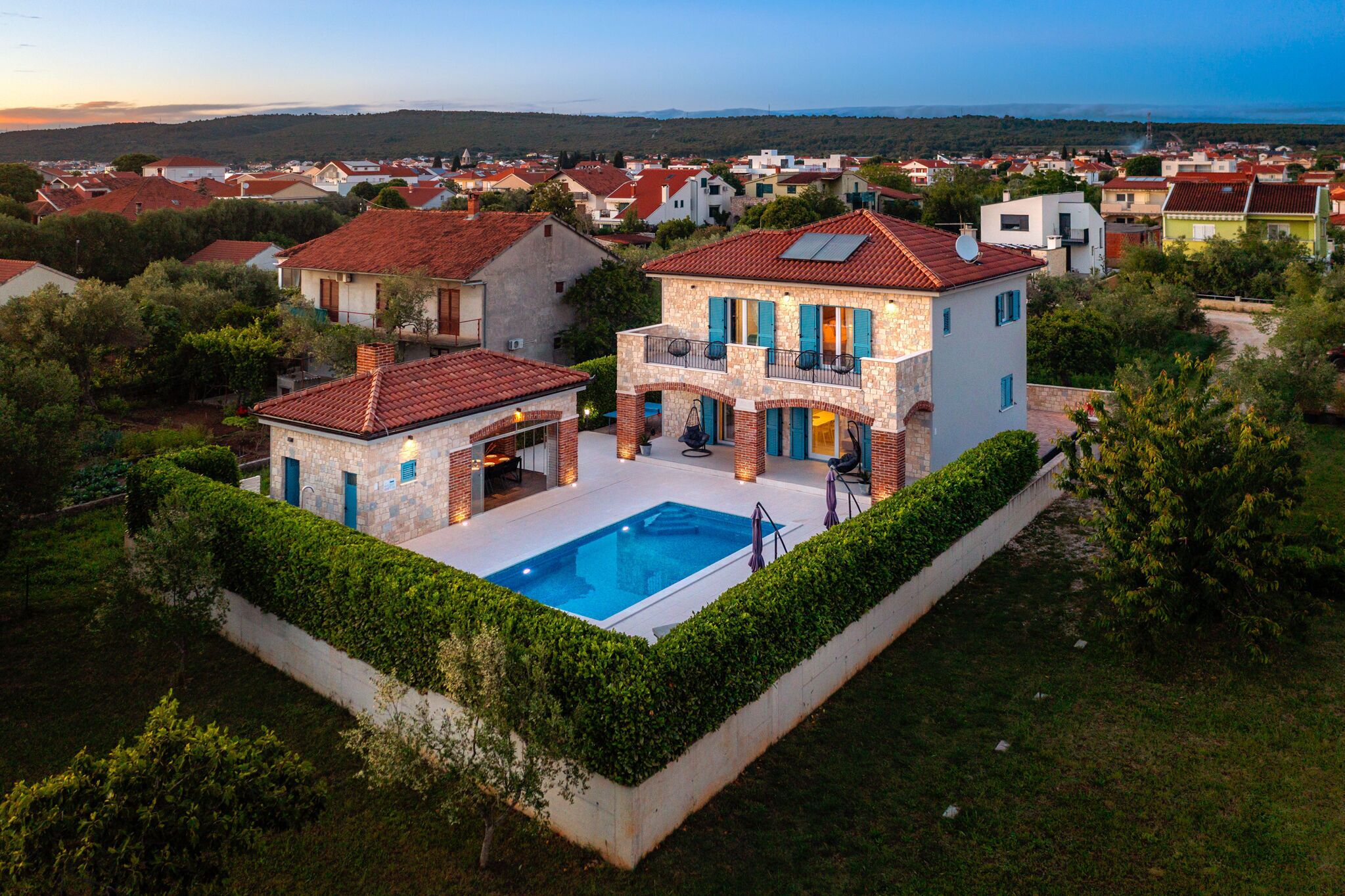 A two-story house with a swimming pool, surrounded by greenery and a view of a residential area.