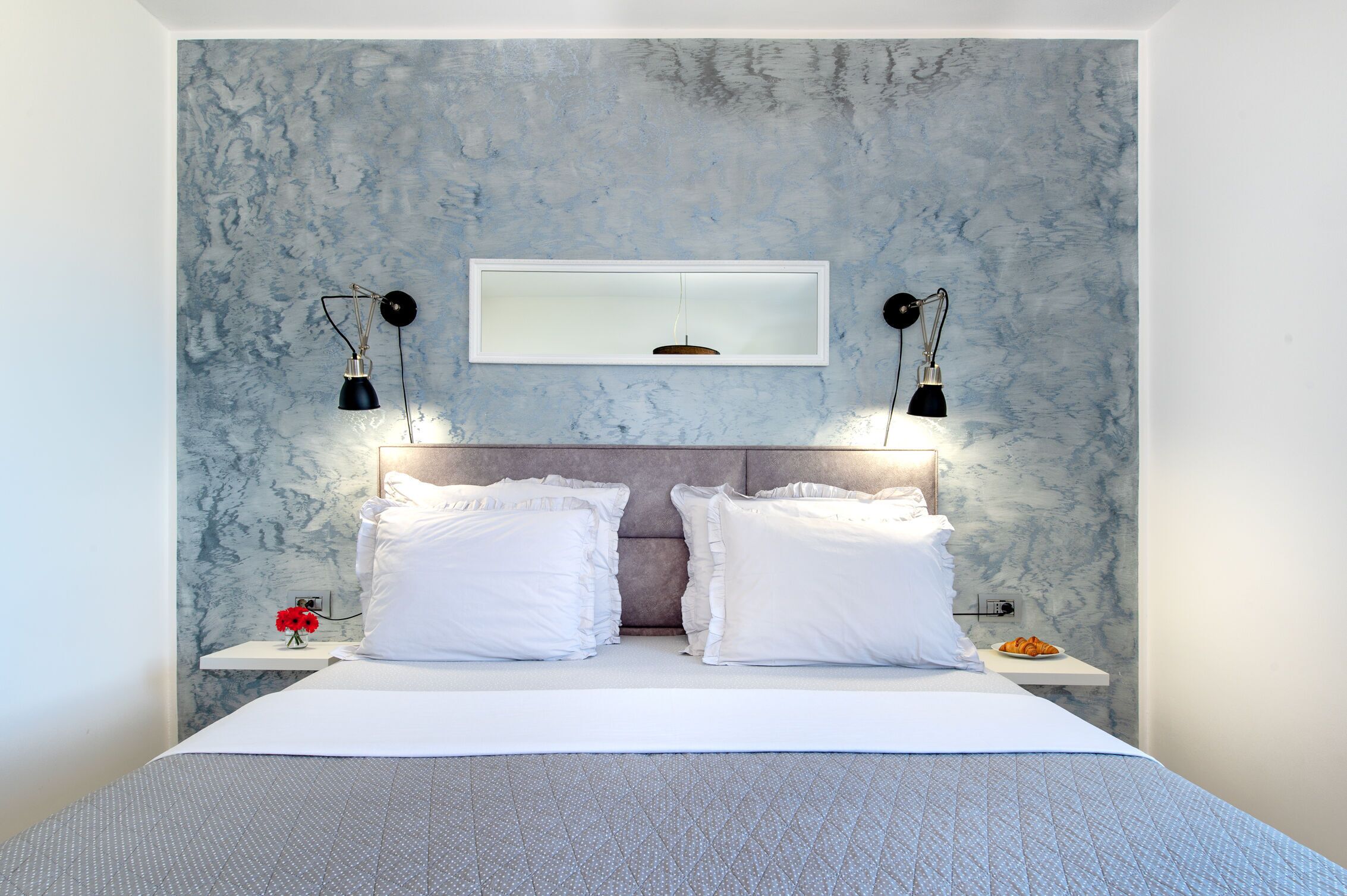 Une chambre moderne avec un mur à motifs de marbre, un lit recouvert de linge de maison blanc, deux tables de chevet avec des lampes et un petit arrangement floral rouge.