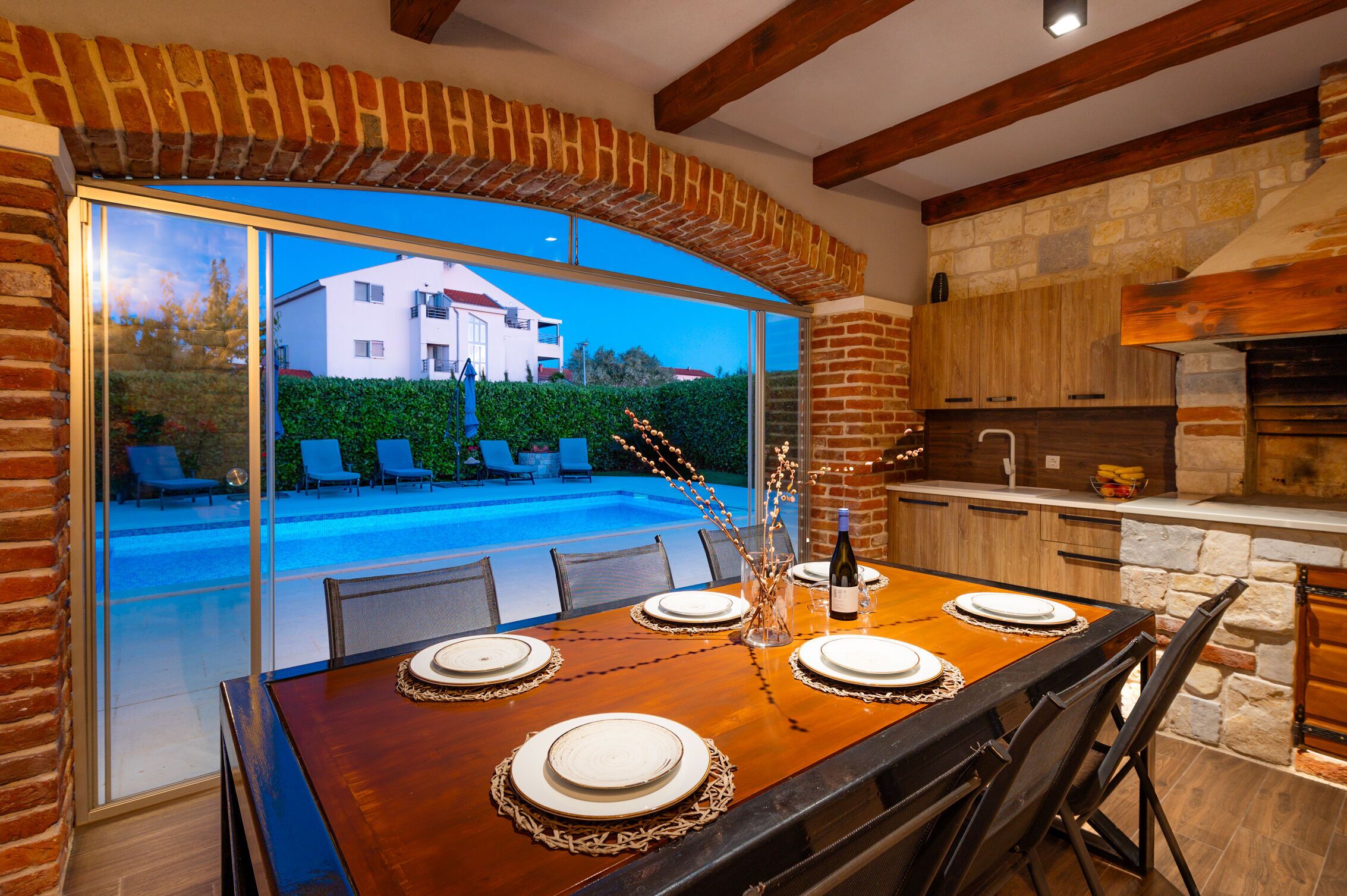 A dining area with a large table set for four, a brick fireplace, and a view of a swimming pool and building outside.