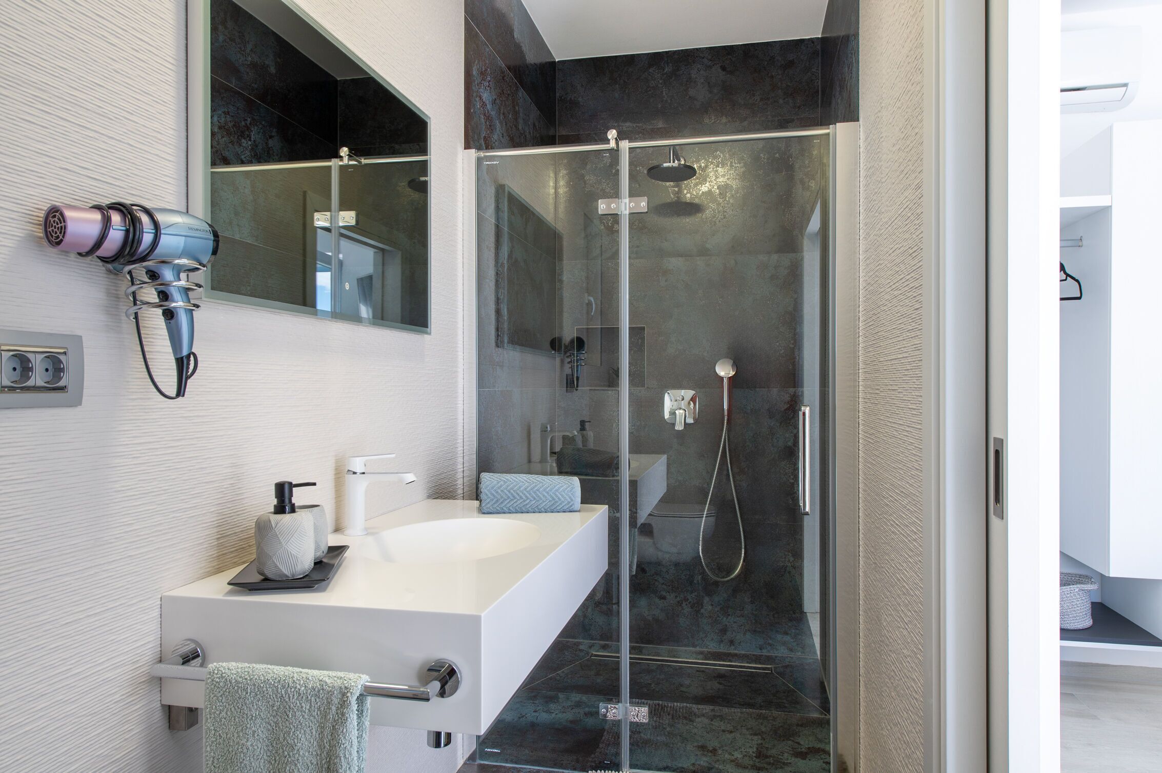 A modern bathroom with a glass-enclosed shower, a white sink, and a hairdryer mounted on the wall.