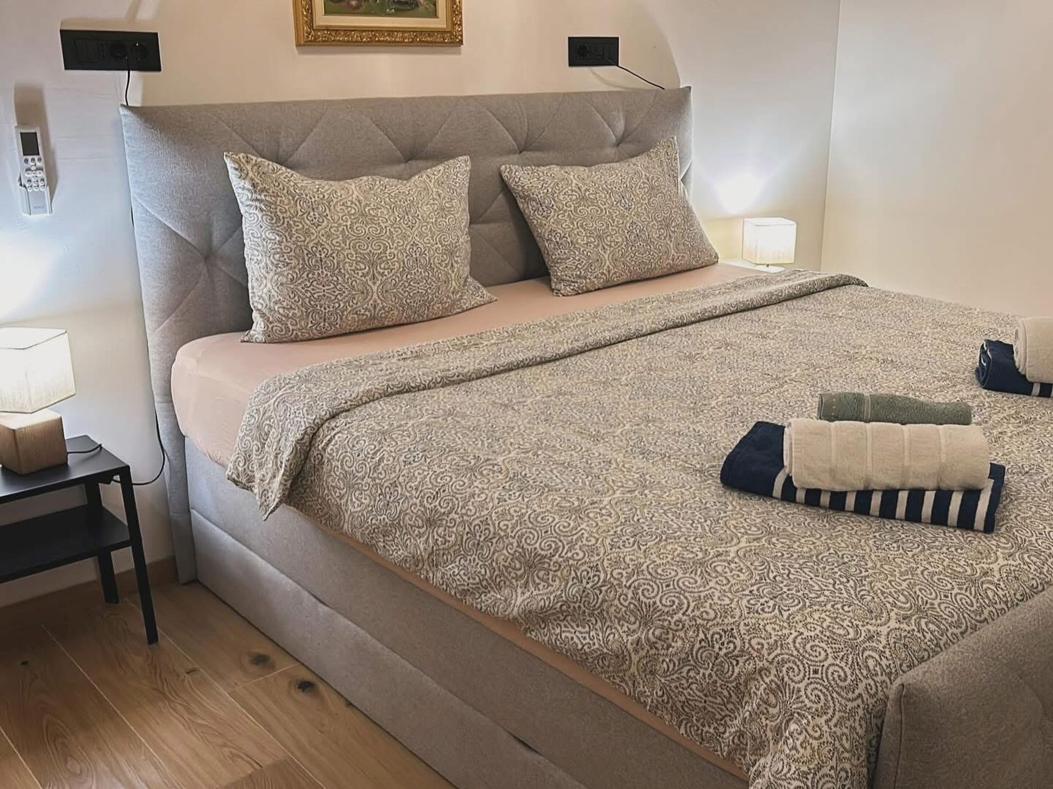 A neatly made bed with patterned bedding, a bedside table with a lamp, and a wall-mounted thermostat.