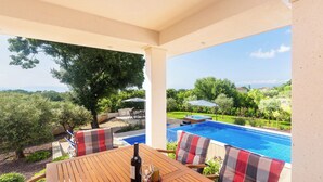 A wooden table with a bottle and glass, outdoor seating with plaid cushions, and a view of a swimming pool and greenery. - Villa Roko (Malinska-Dubasnica)