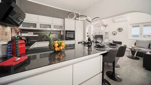 Eine moderne Küche mit einer zentralen Kochinsel, ein Essbereich mit rundem Tisch und ein Wohnbereich mit Sofa und Blick ins Freie.