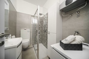 Un bagno moderno con doccia vetrata, un WC bianco e un cesto portasciugamani nero sul lavabo.